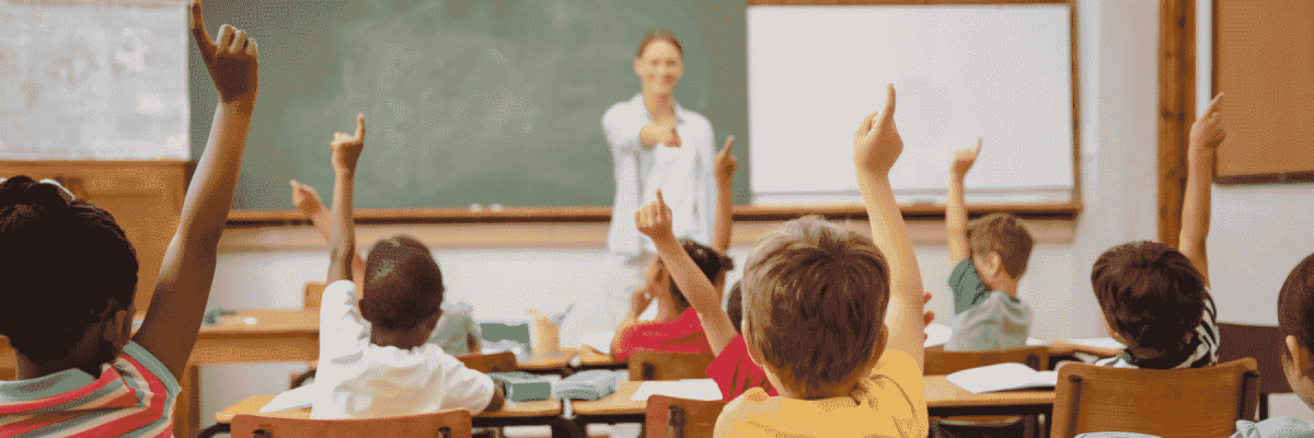 Students raising their hands in a classroom with the teacher at the front