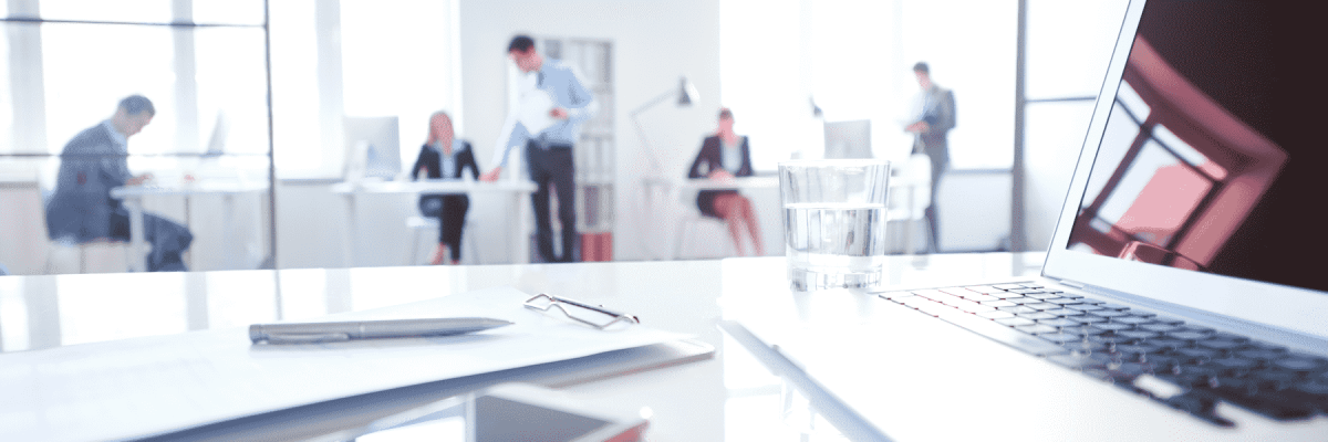 A focused shot of a work desk with a laptop, water cup and notepad with people in the blurred in the background